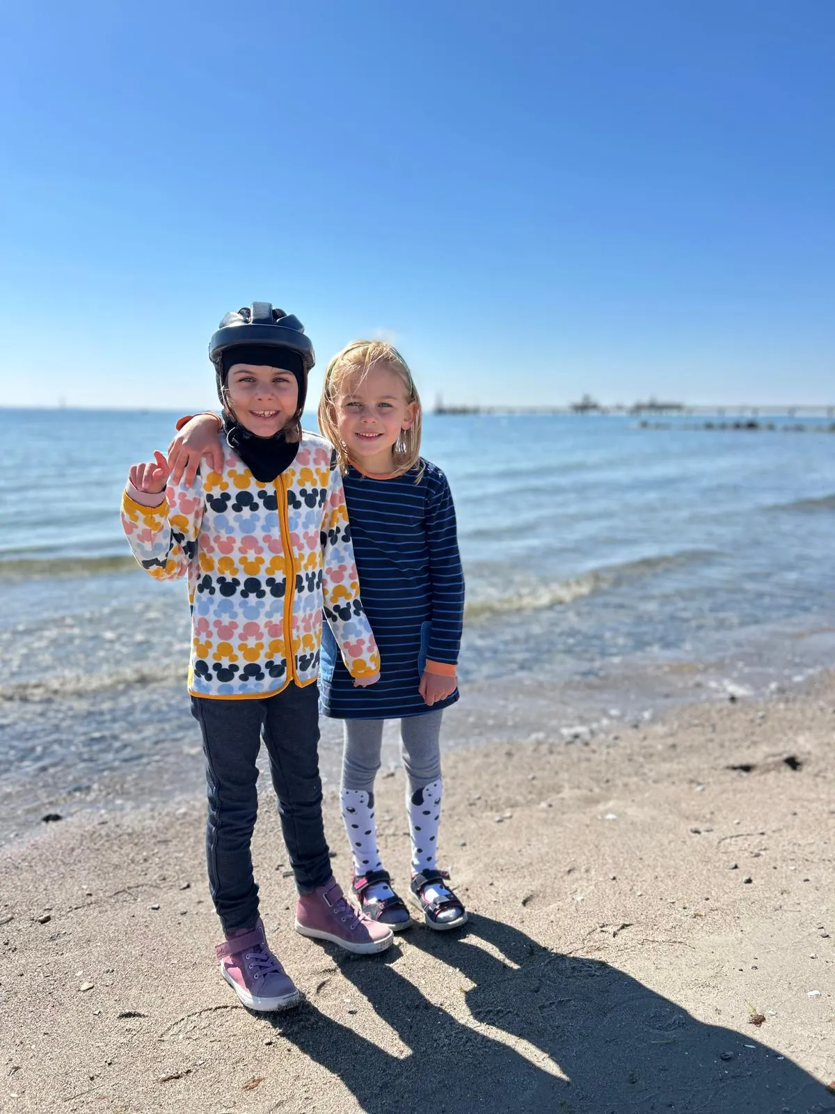 Zwei Mädchen stehen am Strand mit einem Helm und lächeln in die Kamera.
