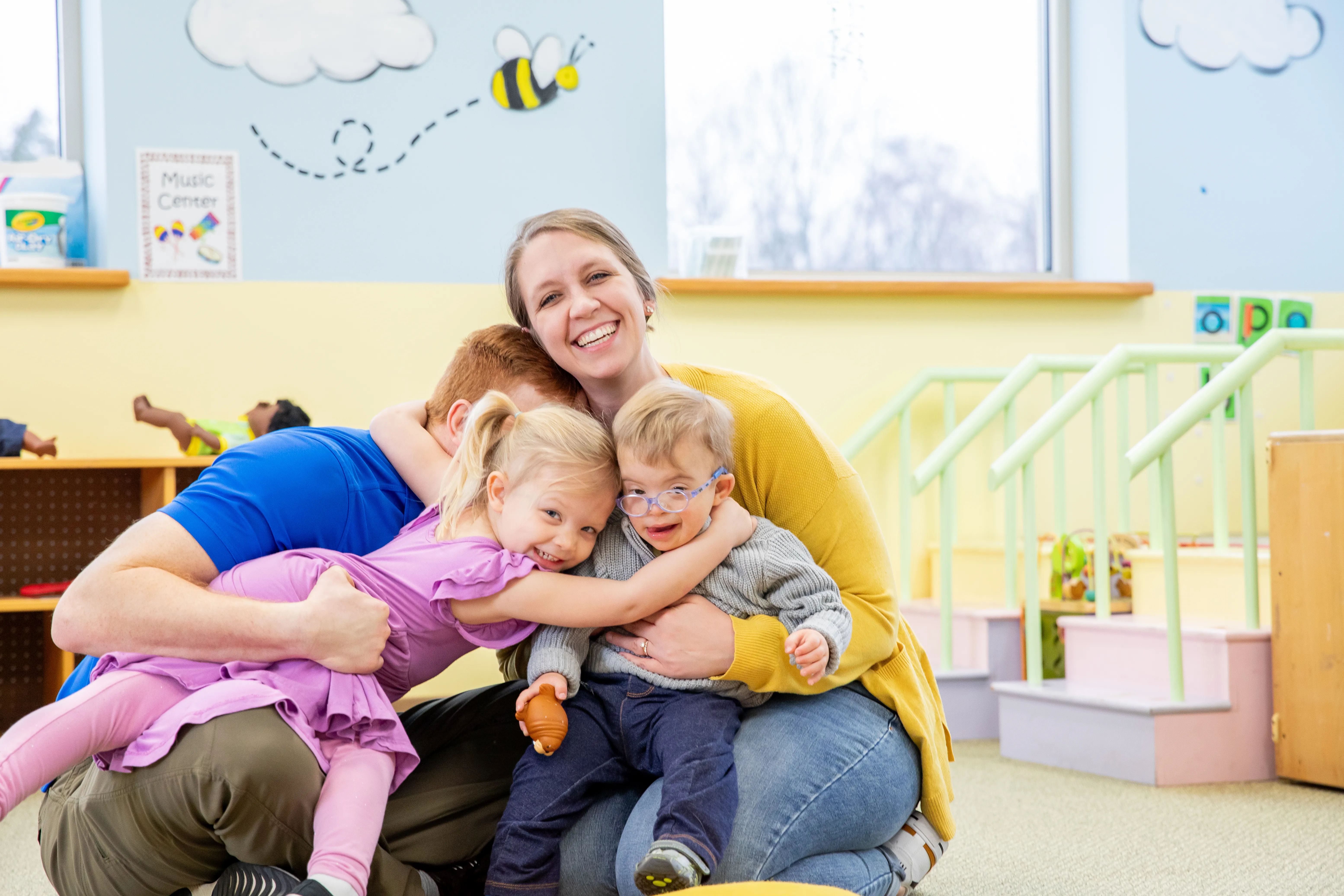 Ein Elternteil umarmt zwei Kinder in einem Spielraum mit bunten Wänden und Spielzeug.