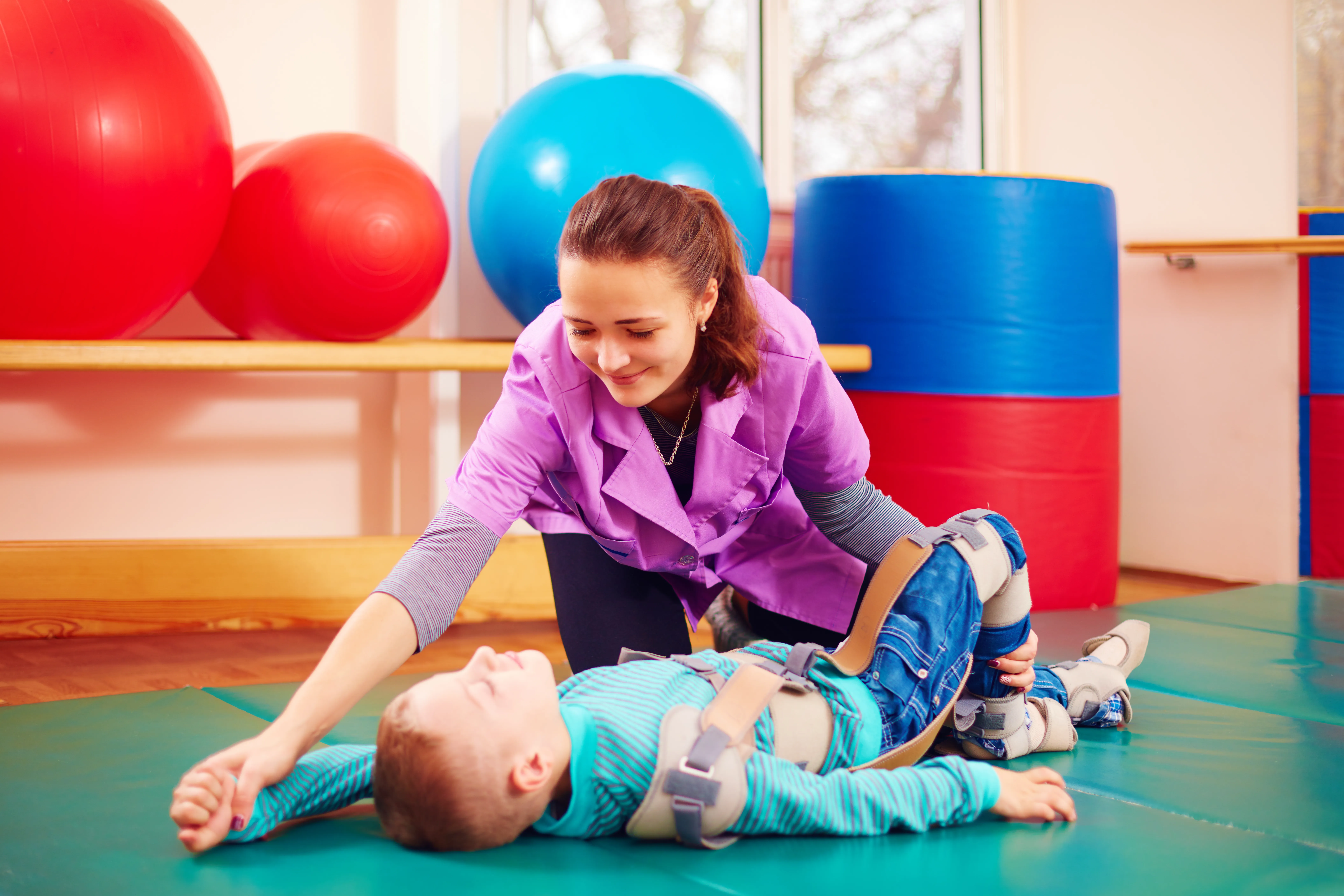 Eine Therapeutin hilft einem Kind, das auf einer Matte liegt, während bunte Fitnessbälle im Hintergrund sichtbar sind.